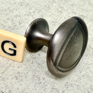 Może przedstawiać: Owalne pokrętło do szafki w kolorze ciemnego brązu z pasującym trzpieniem i podstawą. Obok pokrętła znajduje się kremowa płytka z literą "G". Pokrętło ma szczotkowane metalowe wykończenie.