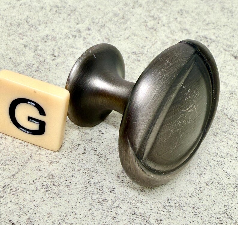 Może przedstawiać: Owalne pokrętło do szafki w kolorze ciemnego brązu z pasującym trzpieniem i podstawą. Obok pokrętła znajduje się kremowa płytka z literą "G". Pokrętło ma szczotkowane metalowe wykończenie.