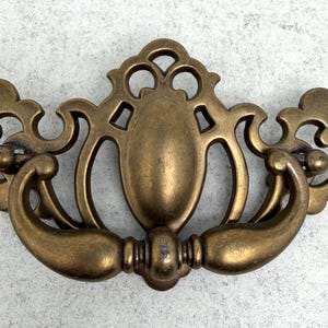 May include: A vintage-style brass drawer pull with an ornate design. The pull features a central oval shape with decorative cutouts and curved handles on either side. The brass has a warm, aged patina, suggesting it is an antique or vintage piece.