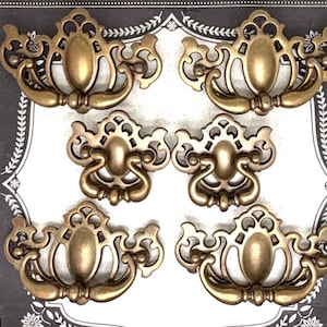 May include: Six antique brass drawer pulls with ornate designs. Each pull features a central oval shape with a curved handle and decorative flourishes. The pulls are arranged on a patterned background.
