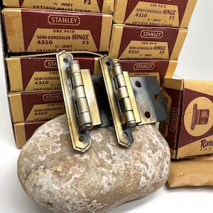 May include: Two antique brass finish Stanley 4310 semi-concealed hinges with inset design. The hinges are on a rock with several boxes of Stanley hinges in the background.