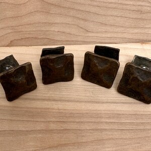 Puede incluir: Cuatro tiradores cuadrados de armario de color bronce con una superficie texturizada. Los tiradores est&aacute;n dispuestos sobre una superficie de madera de color claro, mostrando su apariencia antigua. Estos tiradores son ideales para a&ntilde;adir un toque vintage a los muebles.