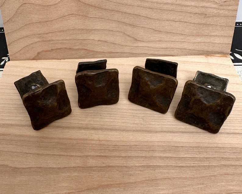 Puede incluir: Cuatro tiradores cuadrados de armario de color bronce con una superficie texturizada. Los tiradores est&aacute;n dispuestos sobre una superficie de madera de color claro, mostrando su apariencia antigua. Estos tiradores son ideales para a&ntilde;adir un toque vintage a los muebles.