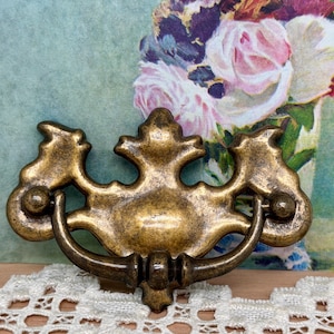 May include: Antique brass-toned drawer pull with a decorative design. The pull features a curved handle and ornate detailing, including stylized floral or leaf-like elements. The hardware is likely for a cabinet or dresser.