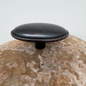 May include: A dark bronze cabinet knob with a rounded, slightly domed top. The knob has a simple, classic design and a small, cylindrical base. The finish has a subtle, aged appearance, adding a touch of vintage charm. Ideal for furniture restoration or new builds.