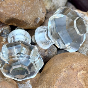 May include: Close-up of several clear, faceted glass cabinet knobs. The knobs have a geometric design and are resting on a bed of smooth, brown rocks. The knobs are reflecting light, creating a sparkling effect. These knobs are ideal for adding a touch of elegance to furniture.