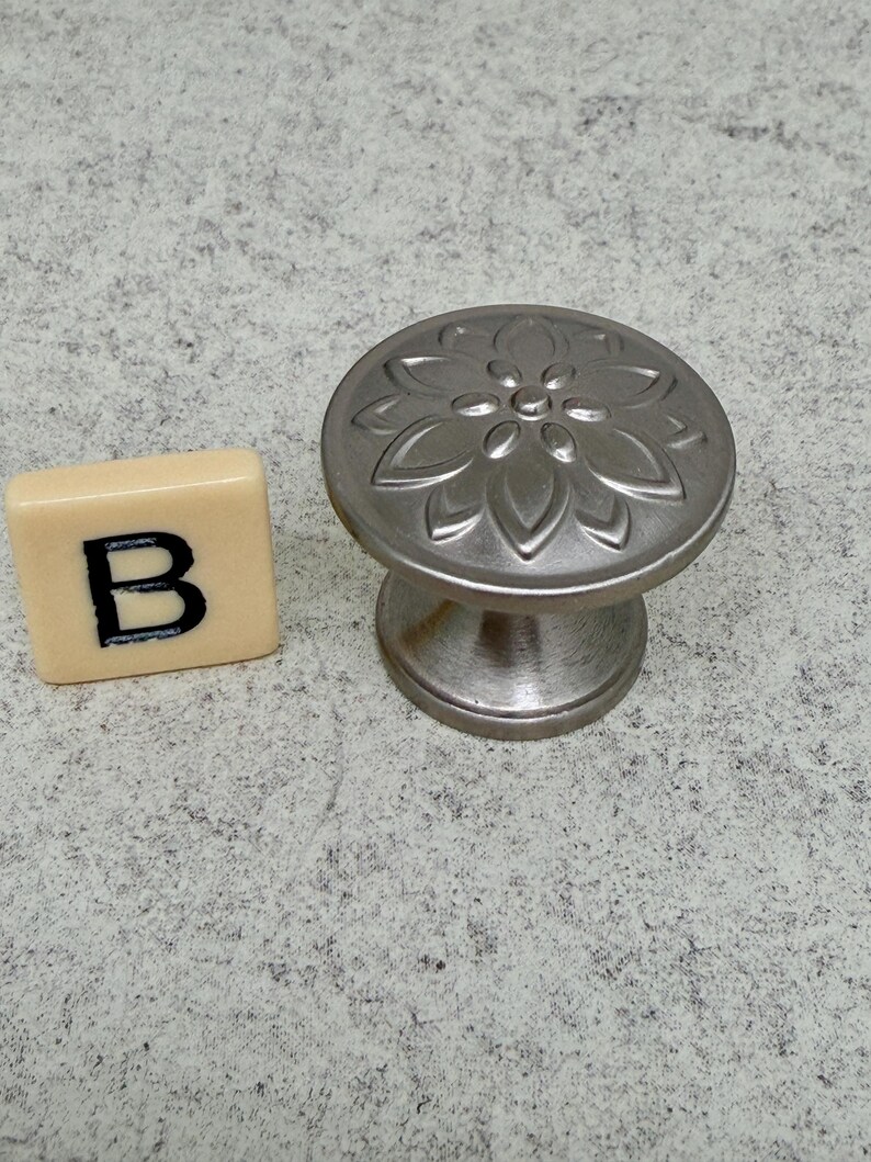 Może przedstawiać: Szczotkowany srebrny uchwyt do szafki z kwiatowym wzorem na okrągłej powierzchni. Uchwyt znajduje się obok kwadratowej płytki z literą "B". Uchwyt ma średnicę około 3,8 cm.