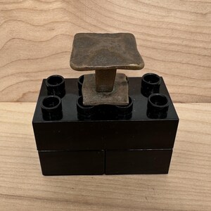 Puede incluir: Una escultura artesanal &uacute;nica con una parte superior cuadrada de color bronce sostenida por un tallo, colocada sobre una base rectangular negra con cuatro aberturas circulares. La pieza se exhibe sobre un fondo de vetas de madera clara.