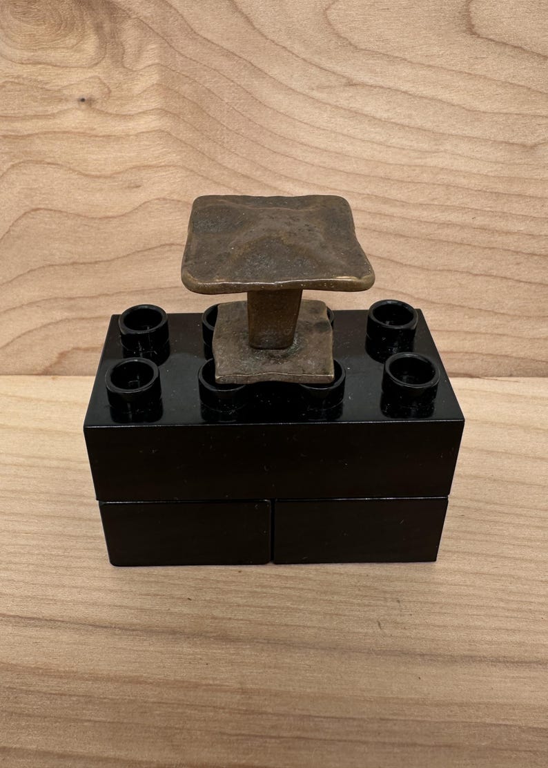 Puede incluir: Una escultura artesanal &uacute;nica con una parte superior cuadrada de color bronce sostenida por un tallo, colocada sobre una base rectangular negra con cuatro aberturas circulares. La pieza se exhibe sobre un fondo de vetas de madera clara.