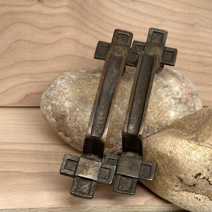 May include: Two antique bronze cabinet pulls with a cross design. The pulls are rectangular with a central handle and cross-shaped ends. The bronze has a weathered, aged appearance, suggesting vintage hardware.