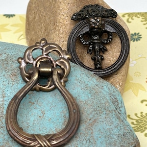 May include: Two antique-style cabinet door pulls. One is a round pull with a decorative floral design and the other is a ring pull with a twisted rope design.
