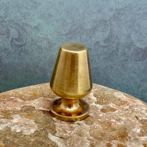 May include: A small, brass-colored lamp finial with a conical shade and a circular base. The finial is made of a shiny, metallic material and is designed to be mounted on a lamp. The overall design is simple and elegant.