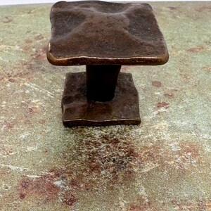 Puede incluir: Un tirador de armario cuadrado de color bronce con una superficie texturizada. El tirador tiene una base cuadrada y una parte superior cuadrada texturizada. El herraje es probablemente para un caj&oacute;n o armario.