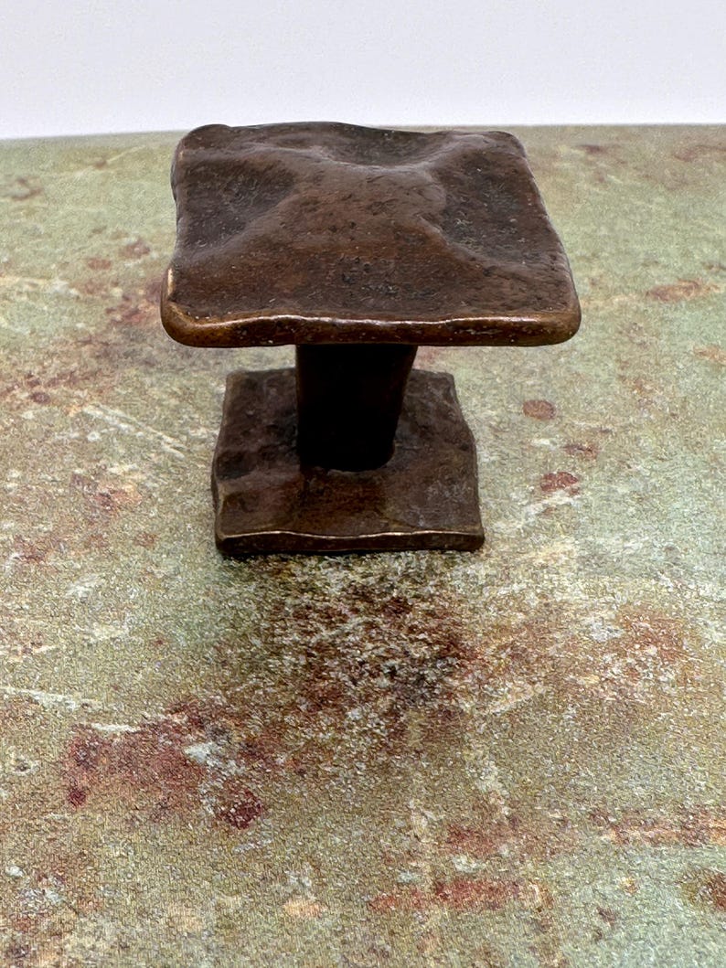 Puede incluir: Un tirador de armario cuadrado de color bronce con una superficie texturizada. El tirador tiene una base cuadrada y una parte superior cuadrada texturizada. El herraje es probablemente para un caj&oacute;n o armario.