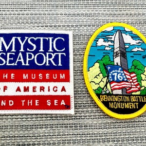 May include: Two embroidered patches. The square patch features blue, white, and red colors with the text MYSTIC SEAPORT THE MUSEUM OF AMERICA AND THE SEA. The oval patch has a yellow border and depicts the Bennington Battle Monument with a 76 flag.