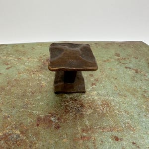 Puede incluir: Un tirador cuadrado de color bronce con una superficie texturizada. El tirador est&aacute; montado sobre una superficie moteada verde y marr&oacute;n. El tirador parece ser de metal, probablemente para un caj&oacute;n o armario.