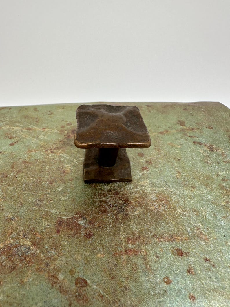 Puede incluir: Un tirador cuadrado de color bronce con una superficie texturizada. El tirador est&aacute; montado sobre una superficie moteada verde y marr&oacute;n. El tirador parece ser de metal, probablemente para un caj&oacute;n o armario.