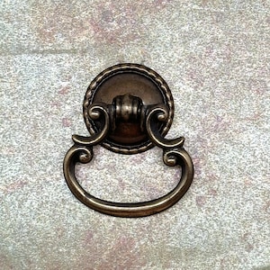 May include: Antique bronze-toned drawer pull with a circular backplate and a decorative, curved handle. The backplate features a rope-like border. The handle has a detailed, ornate design, suitable for furniture restoration or decorative projects.