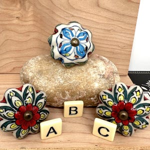 May include: Three decorative ceramic knobs with floral designs. Two knobs have red and green petals with a brass center, and one has blue and white petals. Small lettered tiles spell out "A", "B", and "C".