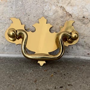 May include: Gold-toned metal cabinet pull with a decorative, ornate design. The pull has a curved handle and two round knobs.