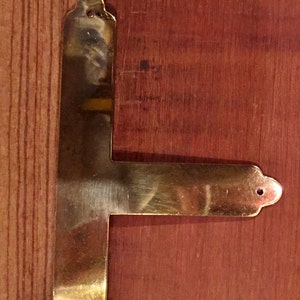 May include: A brass corner brace with a decorative design. The brace is attached to a wooden surface.