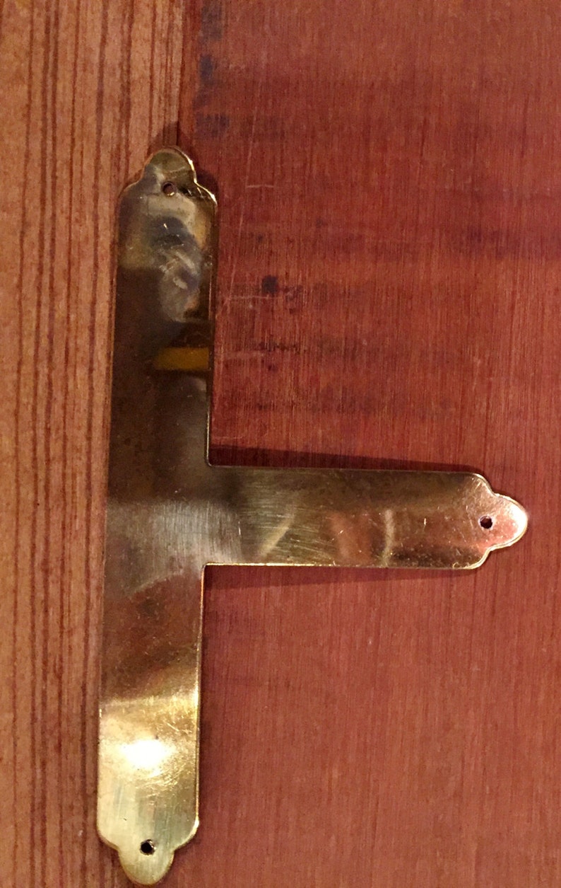 May include: A brass corner brace with a decorative design. The brace is attached to a wooden surface.