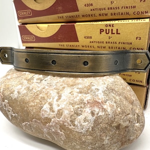 May include: Antique brass door pull with four rivets and a beveled edge. The pull is 5 inches long and is labeled "ONE PULL" on a box with the Stanley Works logo.