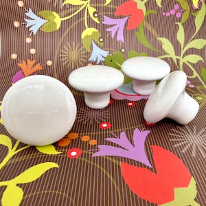 May include: A collection of white ceramic cabinet knobs in various shapes. One is a sphere, and the others are mushroom-shaped. The knobs are arranged on a brown background with a colorful floral pattern. These knobs are ideal for adding a touch of elegance to furniture.