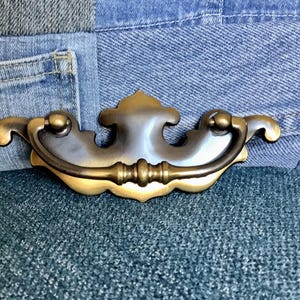 May include: A brass drawer pull with a decorative, ornate design. The pull has a curved handle and a decorative, scalloped edge.