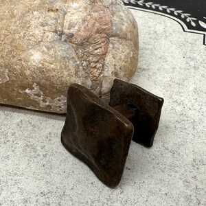 Puede incluir: Dos tiradores de armario cuadrados de color marr&oacute;n oscuro con una superficie texturizada. Los tiradores est&aacute;n hechos de un material met&aacute;lico y tienen un aspecto r&uacute;stico y antiguo. Una piedra grande y de color claro est&aacute; en el fondo.