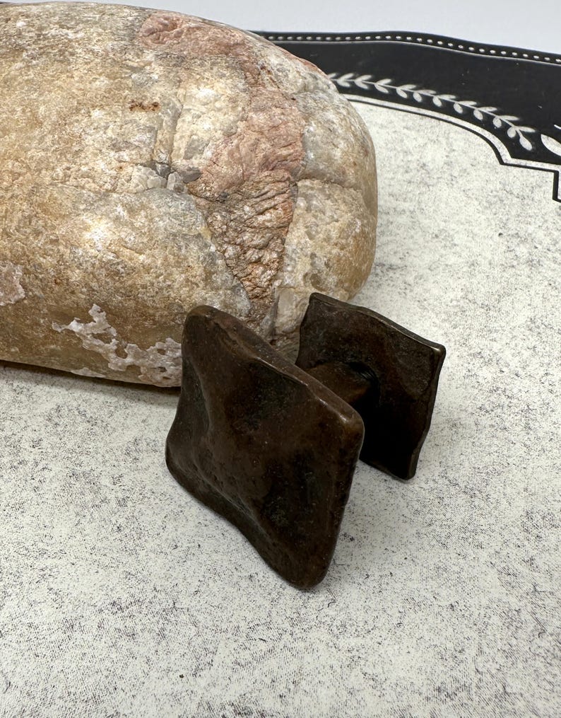 Puede incluir: Dos tiradores de armario cuadrados de color marr&oacute;n oscuro con una superficie texturizada. Los tiradores est&aacute;n hechos de un material met&aacute;lico y tienen un aspecto r&uacute;stico y antiguo. Una piedra grande y de color claro est&aacute; en el fondo.