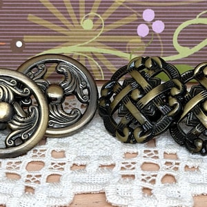 May include: Close-up of decorative bronze-colored cabinet knobs. Two knobs have an ornate, floral design, and two have a woven pattern. The letter "A" and "B" are on small, off-white tiles. The knobs are on a white lace surface.