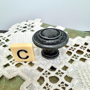 Może przedstawiać: Gałka do szafki z postarzanego metalu w kolorze ciemnoszarym ze srebrnymi akcentami i okrągłym wzorem. Obok gałki znajduje się płytka z literą "C". Gałka jest prawdopodobnie przeznaczona do mebli lub dekoracji wnętrz.