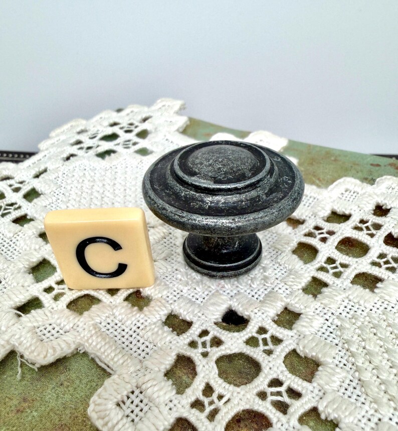 Może przedstawiać: Gałka do szafki z postarzanego metalu w kolorze ciemnoszarym ze srebrnymi akcentami i okrągłym wzorem. Obok gałki znajduje się płytka z literą "C". Gałka jest prawdopodobnie przeznaczona do mebli lub dekoracji wnętrz.