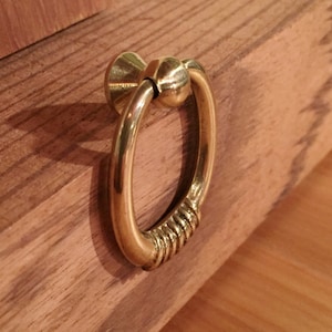 May include: A gold-toned ring pull cabinet handle with a round knob. The handle is attached to a wooden surface.