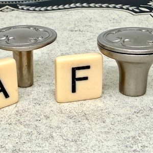 Może przedstawiać: Dwa srebrne gałki do szafek z dekoracyjnymi detalami i dwie kremowe płytki z literami "A" i "F" w kolorze czarnym. Gałki mają kształt grzyba, odpowiednie do mebli.