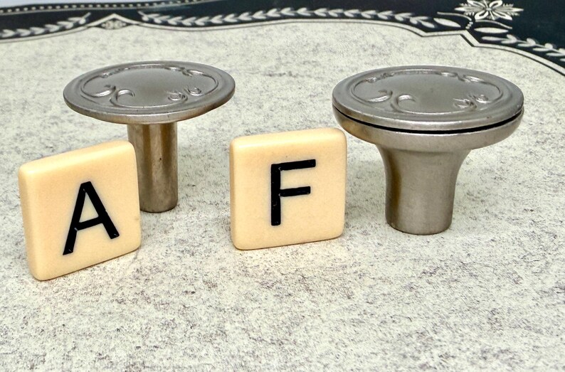 Może przedstawiać: Dwa srebrne gałki do szafek z dekoracyjnymi detalami i dwie kremowe płytki z literami "A" i "F" w kolorze czarnym. Gałki mają kształt grzyba, odpowiednie do mebli.