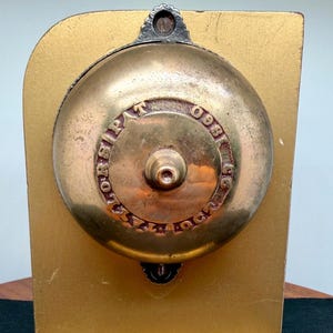 May include: A circular brass bell mounted on a gold-colored rectangular base. The bell features a central knob and embossed text reading TAYLOR'S PAT OCT 93. The metal surface shows signs of age and patina, with a dark metal mounting bracket.