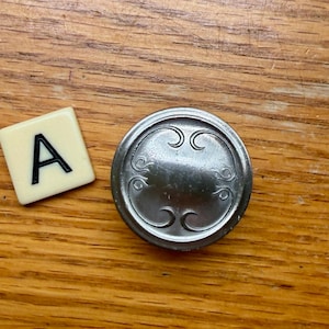 Może przedstawiać: Płytka do Scrabble z literą "A" obok srebrnego, okrągłego guzika z dekoracyjnymi zawijasami. Obiekty znajdują się na drewnianej powierzchni.