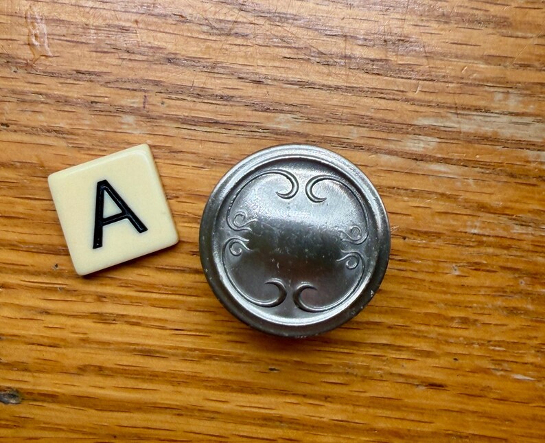 Może przedstawiać: Płytka do Scrabble z literą "A" obok srebrnego, okrągłego guzika z dekoracyjnymi zawijasami. Obiekty znajdują się na drewnianej powierzchni.