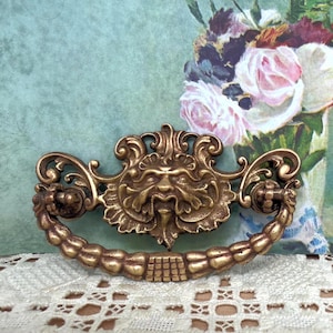 May include: Ornate antique brass drawer pull with a central face design and curved handle. The brass hardware features intricate detailing and a warm, aged finish, perfect for vintage furniture restoration projects.