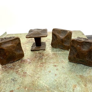 Puede incluir: Cuatro tiradores de armario de color bronce con un dise&ntilde;o cuadrado y texturizado. Un tirador est&aacute; montado sobre un poste corto de color marr&oacute;n oscuro. Los tiradores est&aacute;n dispuestos sobre una superficie moteada verde y marr&oacute;n, con un fondo blanco.