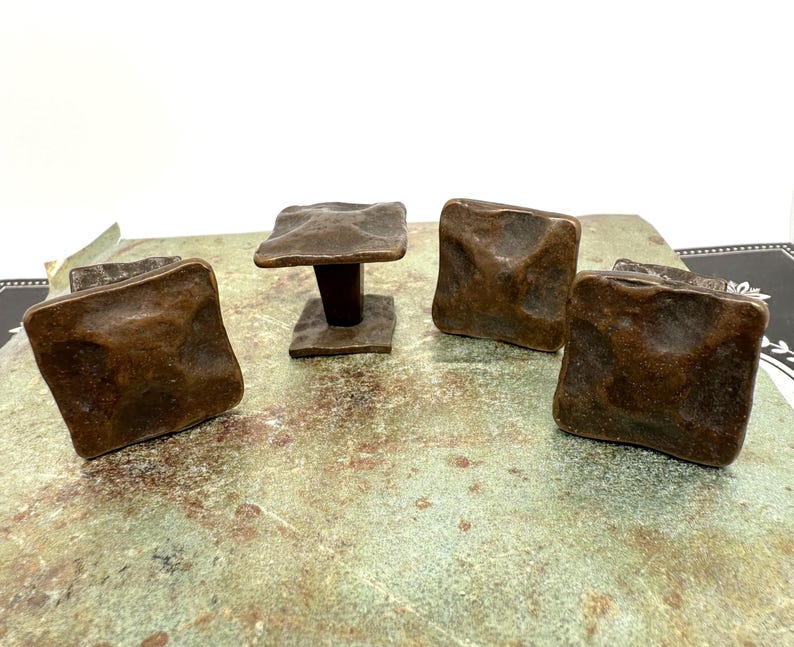 Puede incluir: Cuatro tiradores de armario de color bronce con un dise&ntilde;o cuadrado y texturizado. Un tirador est&aacute; montado sobre un poste corto de color marr&oacute;n oscuro. Los tiradores est&aacute;n dispuestos sobre una superficie moteada verde y marr&oacute;n, con un fondo blanco.