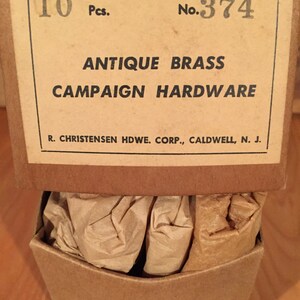 May include: A cardboard box with a label that reads "10 Pcs. No. 374 ANTIQUE BRASS CAMPAIGN HARDWARE R. CHRISTENSEN HDWE. CORP., CALDWELL, N. J.". The box contains brown paper packaging.