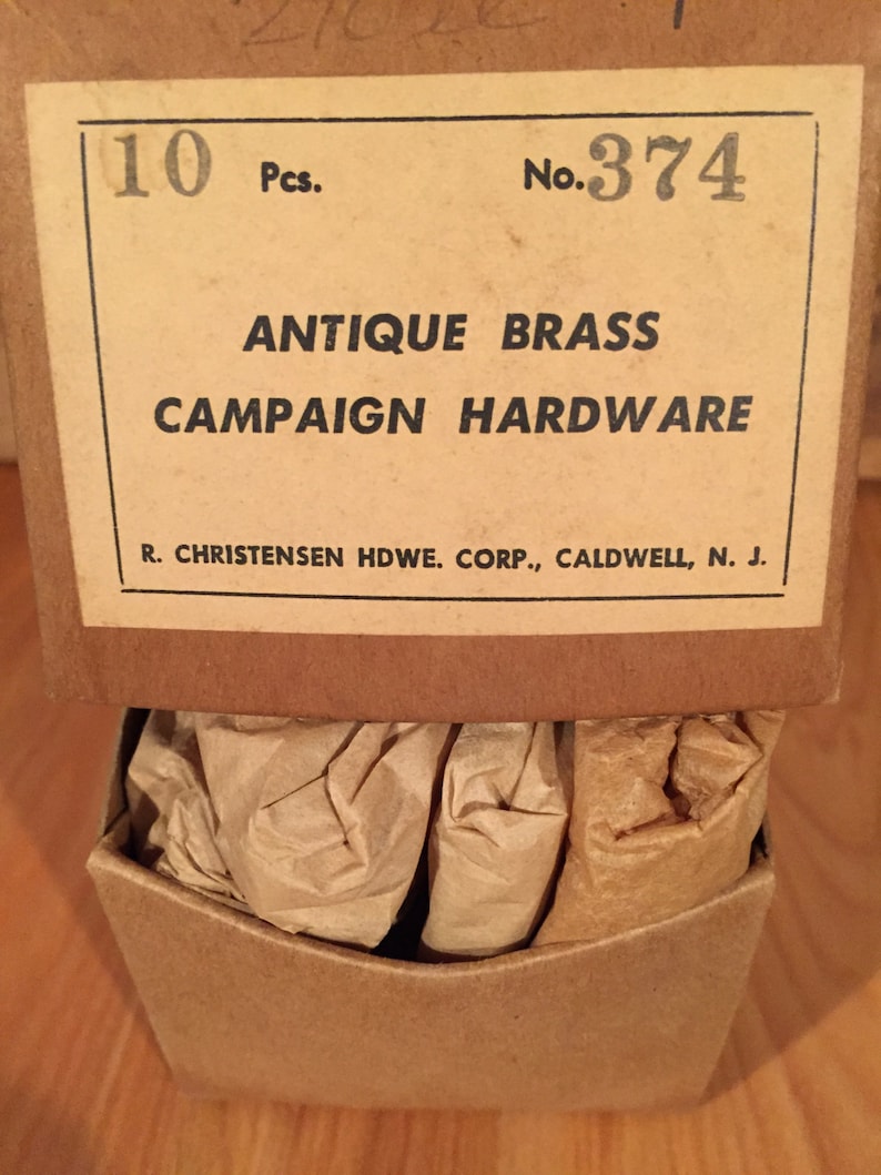 May include: A cardboard box with a label that reads "10 Pcs. No. 374 ANTIQUE BRASS CAMPAIGN HARDWARE R. CHRISTENSEN HDWE. CORP., CALDWELL, N. J.". The box contains brown paper packaging.