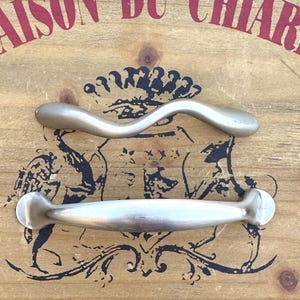 May include: Two silver cabinet door pulls. The top pull is a wavy design. The bottom pull is a straight, rectangular design.