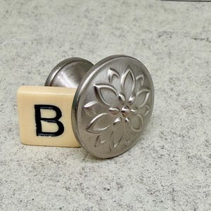 Może przedstawiać: Srebrny uchwyt do szafki z kwiatowym wzorem, połączony z kremową płytką z czarną literą "B". Gałka ma okrągłą, teksturowaną powierzchnię i zakrzywioną podstawę.