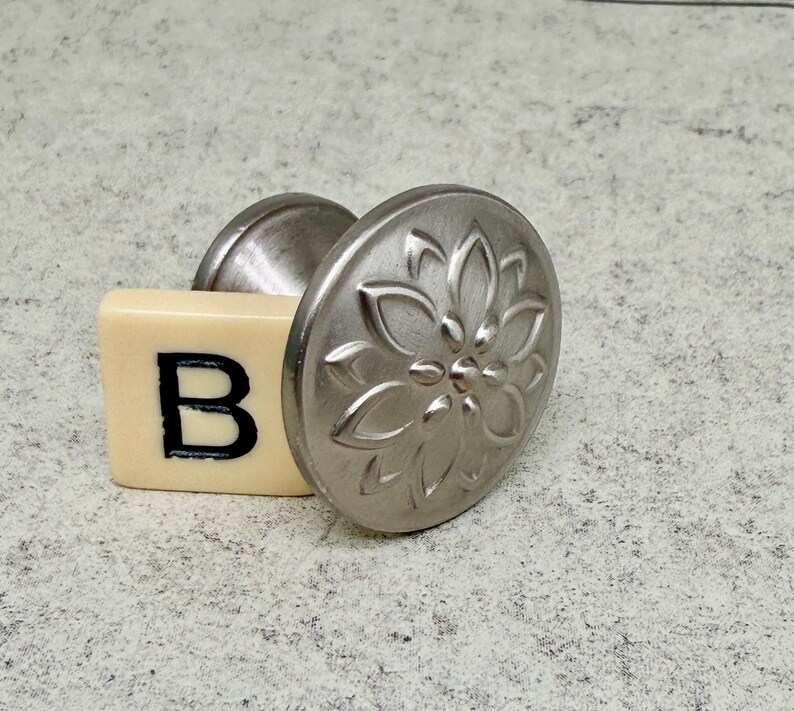 Może przedstawiać: Srebrny uchwyt do szafki z kwiatowym wzorem, połączony z kremową płytką z czarną literą "B". Gałka ma okrągłą, teksturowaną powierzchnię i zakrzywioną podstawę.