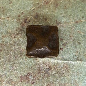 Puede incluir: Un objeto met&aacute;lico cuadrado de color bronce con bordes redondeados. La superficie tiene un aspecto ligeramente texturizado. El objeto est&aacute; sobre un fondo moteado verde y marr&oacute;n.