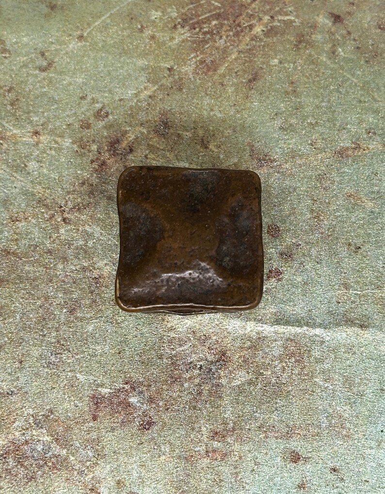 Puede incluir: Un objeto met&aacute;lico cuadrado de color bronce con bordes redondeados. La superficie tiene un aspecto ligeramente texturizado. El objeto est&aacute; sobre un fondo moteado verde y marr&oacute;n.